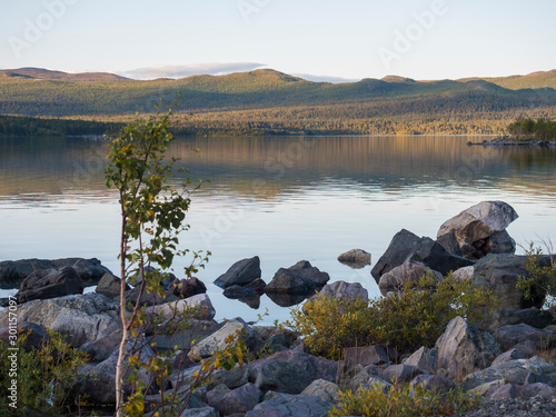 View on river Lulealven in Saltoluokta in Sweden Lapland during sunset golden hour. Green mountain, birch trees, rock boulders clouds and sky and clear the water. photo