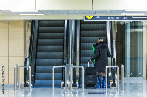 The alone woman on the escalator or moving staircase with inscription departure in English and Chinese in the international airport or railway station from the back moving upstairs with luggage