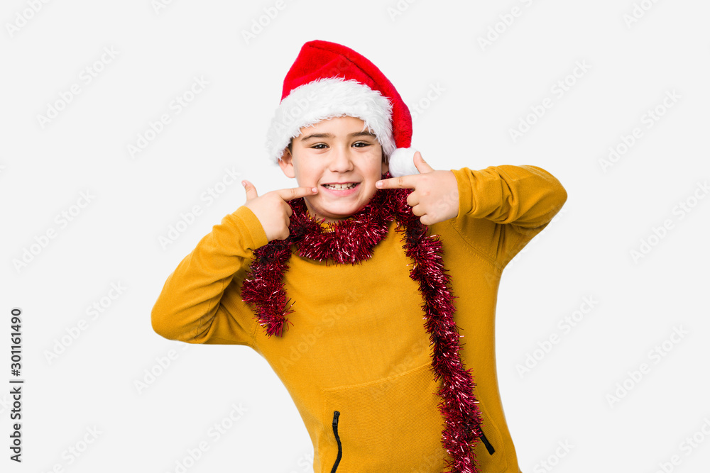 Little boy celebrating christmas day wearing a santa hat isolated smiles, pointing fingers at mouth. Little boy celebrating christmas day wearing a santa hat isolated smiles, pointing fingers at mouth.