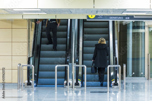 The alone woman on the escalator or moving staircase with inscription departure in English and Chinese in the international airport or railway station from the back moving upstairs with luggage