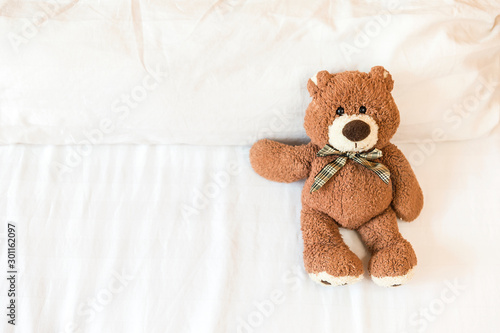 Fluffy brown teddy bear lying down on the bed alone with space on white bed sheet