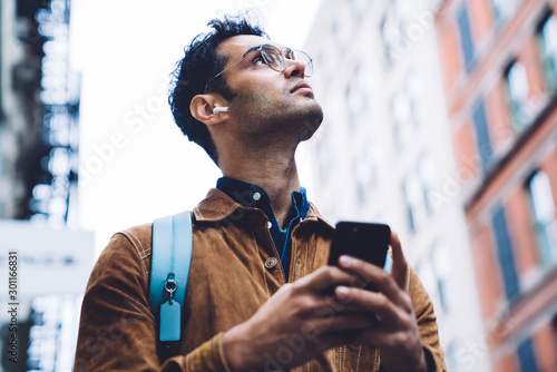 Hispanic young man in eyeglasses standing on street with smartphone