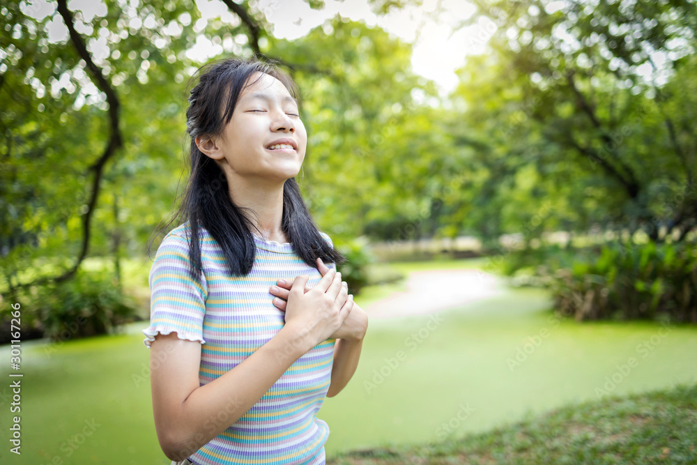 Happy smiling asian child girl standing in green nature,holding hands on heart,female teenage touching her chest,enjoy breathing fresh air with closed eyes,relaxing in park,healthy lifestyle concept Happy smiling asian child girl standing in green nature,holding hands on heart,female teenage touching her chest,enjoy breathing fresh air with closed eyes,relaxing in park,healthy lifestyle concept