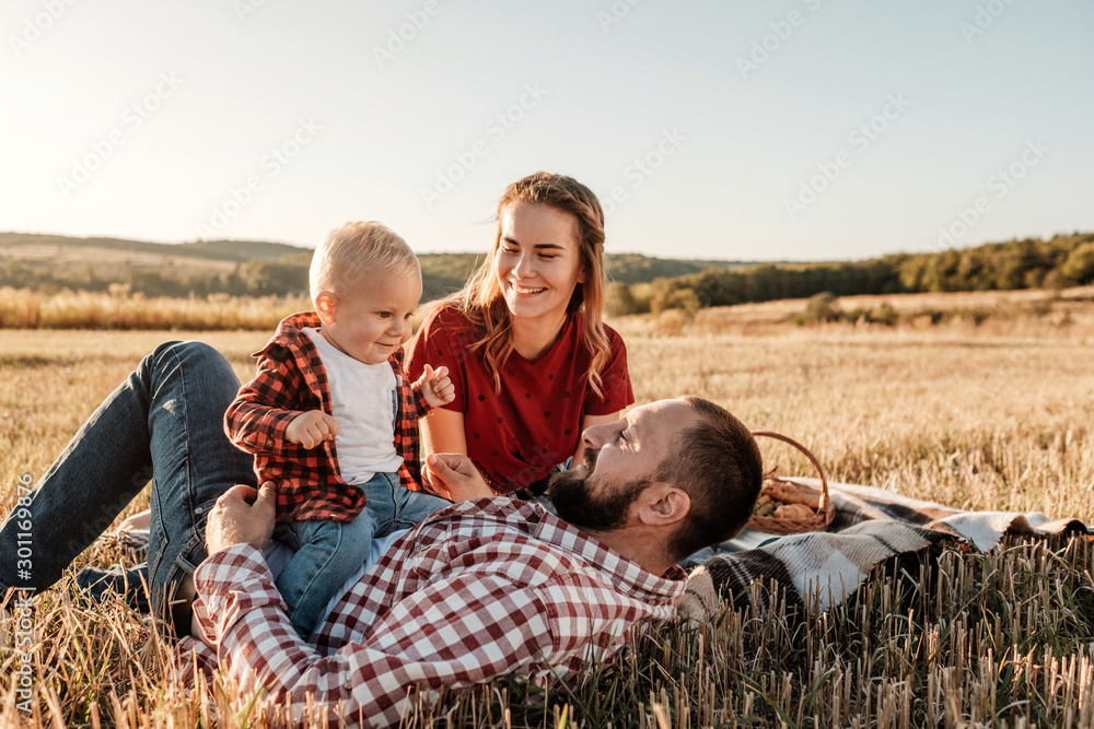 Happy Young Family Mom and Dad with Their Little Son Enjoying Summer Weekend Picnic Outside the City in the Field at Sunny Day Sunset, Vacation Time Concept Happy Young Family Mom and Dad with Their Little Son Enjoying Summer Weekend Picnic Outside the City in the Field at Sunny Day Sunset, Vacation Time Concept