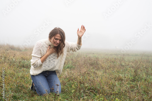 Girl closed her eyes on the knees, praying in a field during beautiful fog. 