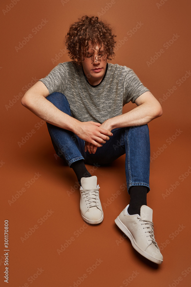Full shot of a curly haired European man in blue jeans, gray tee, gray  sneakers and black socks. The man is posing on the brown background. Stock  Photo | Adobe Stock