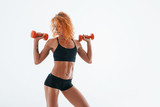 Exercising with red dumbbells. Redhead female bodybuilder is in the studio on white background