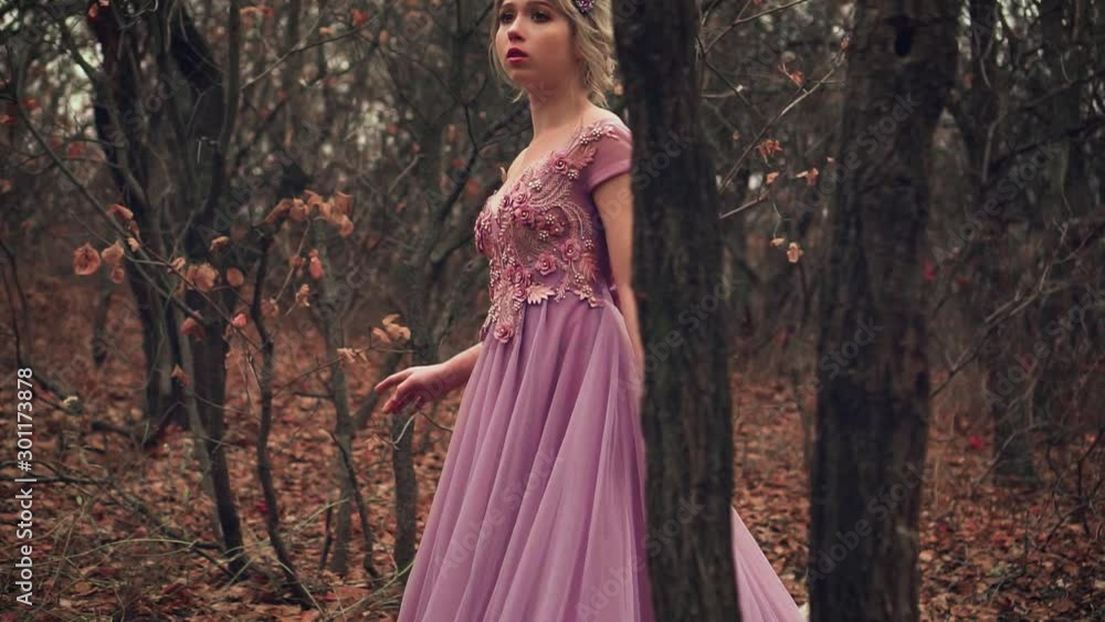 beauty blonde princess is standing in purple lush dress in a corset. Frightened, lost in the forest. Glamorous fashion woman queen in crown. Autumn backdrop, black trees orange leaves. Cute young face