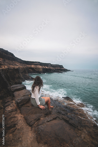 Mujer sentada en acantilado mirando al horizonte con mar océano verde azul en paisaje de montaña y rocas photo