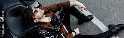 panoramic shot of young man resting while leaning on balck motorcycle and sitting on alphalt