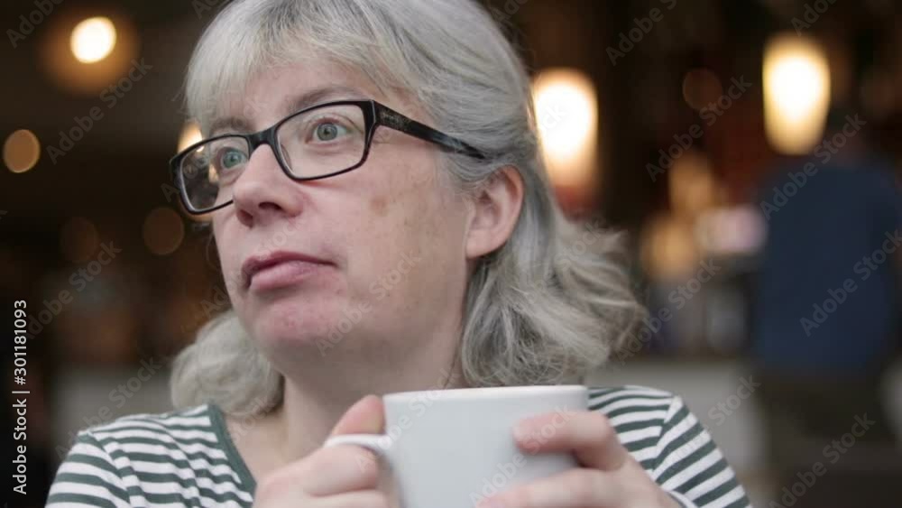 Real Modern Grey Haired, Glasses Wearing, Middle Aged White Female Drinking Coffee In Coffee House, Laughing, Inquisitive, Bokeh Background. Real Modern Grey Haired, Glasses Wearing, Middle Aged White Female Drinking Coffee In Coffee House, Laughing, Inquisitive, Bokeh Background.