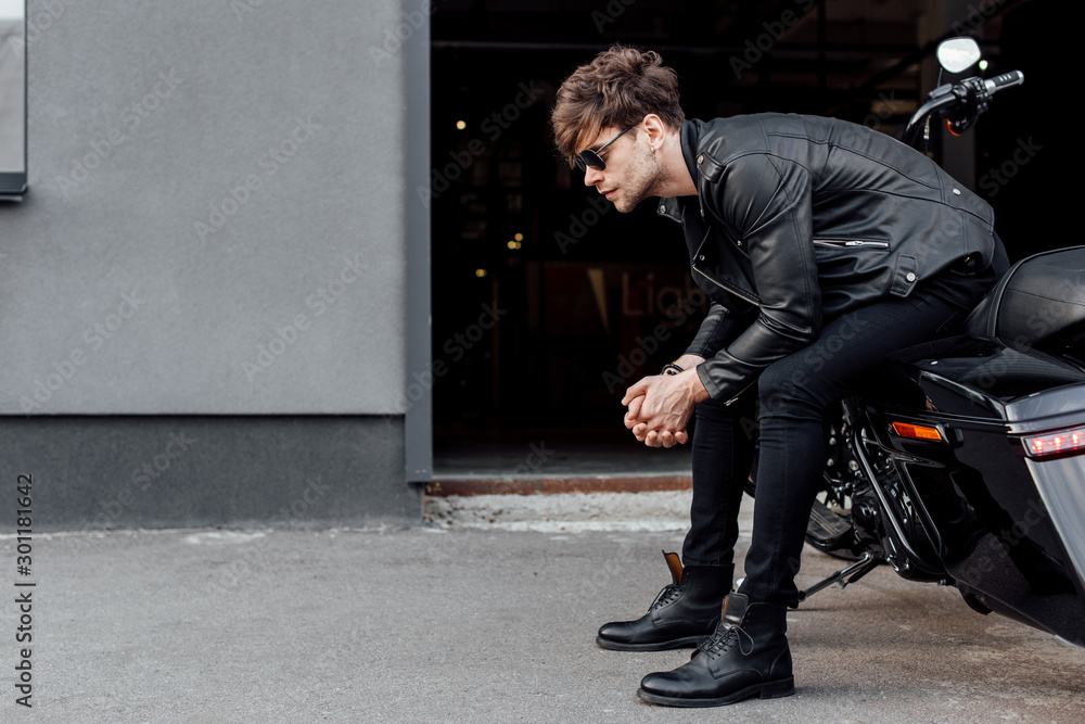 side view of handsome man in leather jacket and sunglasses sitting on black motorcycle and looking forward