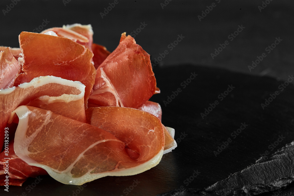 Gourmet, manually sliced jamon on black stone slate board against a dark grey background. Close-up shot. Side view. Copy space.