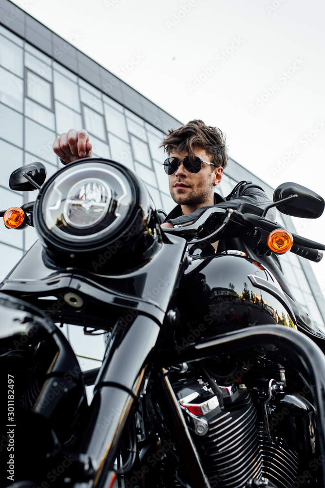 low angle view of handsome young man in sunglasses and leather jacket looking away while sitting on motorcycle