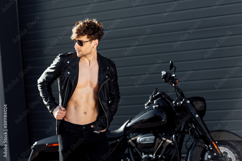 Naklejka premium young man with muscular torso in leather jacket standing near black motorcycle