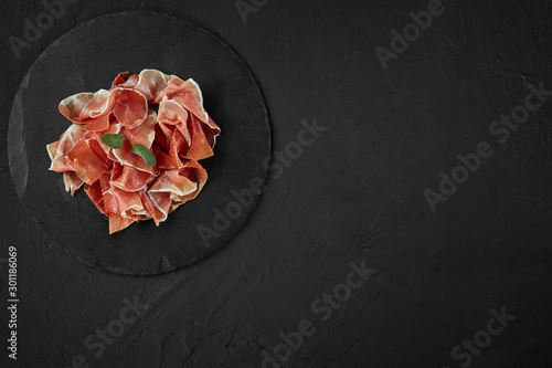 Gourmet, manually sliced jamon with herbs on black stone slate board against a dark grey background. Close-up shot. Top view. Copy space.