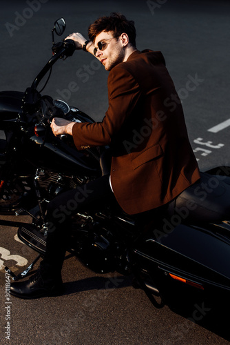 motorcyclist in brown jacket and sunglasses sitting on motorcycle and holding handlebars