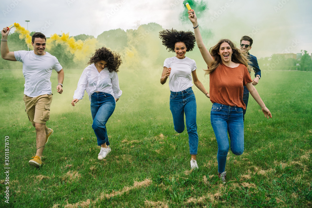 Group of five friends runs in a park with two smoke bombs at the park - Millennials have fun together in the summer at sunset - obrazy, fototapety, plakaty 