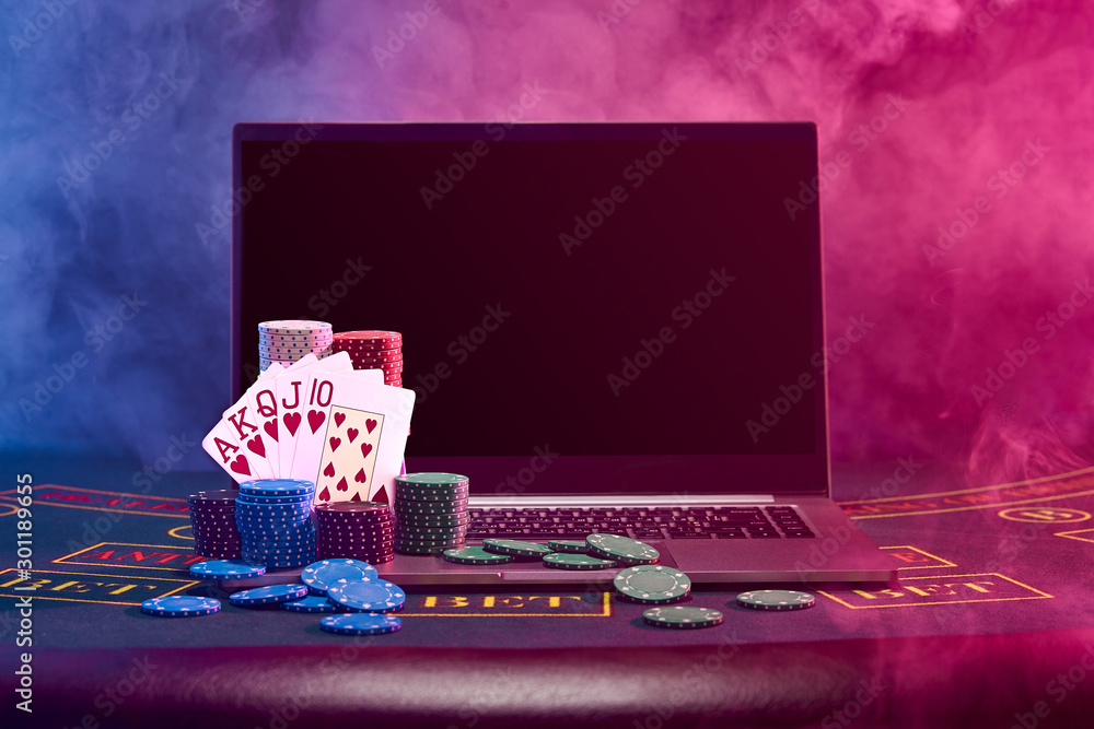 Chips piles and playing cards on laptop standing on blue cover of a table. Black, smoke background. Gambling, poker, casino concept. Close-up.