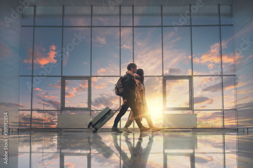 Picture of young couple walking in aurport close to window. Sunset outside. Going for a trip or vacation. Travel together. Take suitcase and backpacks.