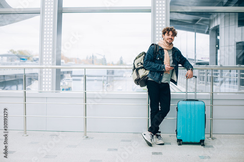 Young handsome happy man stand outside and wait. Hold hand on suitcase. Carry backpack on back. Waiting for flight. Pose on camera and smile. Hold phone in hands.