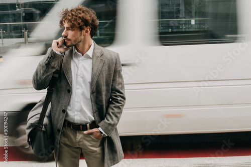 Careful young businessman walking on crosswalk and look to left. Talk on phone. Car riding behind. Blurred background. Posing on camera. Hold purse on shoulder.