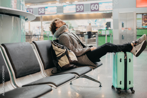 Young businessman sleep alone in waiting room. Sit in airport or train station. Hold legs on suitcase. Backpack besides. Waiting for flight or train. Expert manager travel abroad. Business trip.