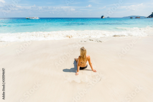 Blonde young woman in black swimsuit lying on side at beach. Beautiful tanned girl lying on sand. Attractive girl wearing glamour swimwear on tropical beach while sunbathing.