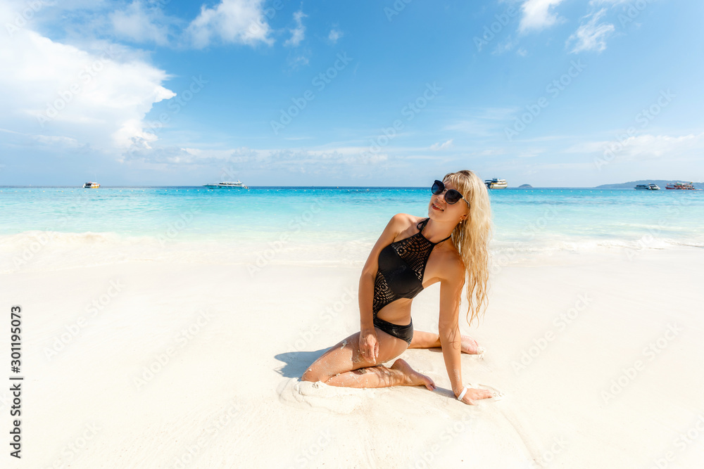 Blonde young woman in black swimsuit sitting on side at beach. Beautiful tanned girl lying on sand. Attractive girl wearing glamour swimwear on tropical beach while sunbathing