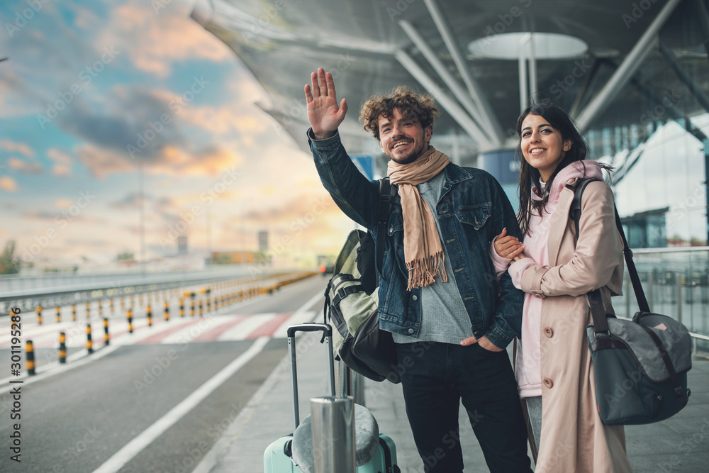 Young man and woman stand outside airport and wait for taxi cab. Guy wave with hand. After vacation or travel. Georgian woman hold his hand and smile. Sunset outside. Evening. - obrazy, fototapety, plakaty 