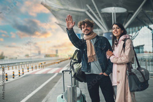 Young man and woman stand outside airport and wait for taxi cab. Guy wave with hand. After vacation or travel. Georgian woman hold his hand and smile. Sunset outside. Evening. photo