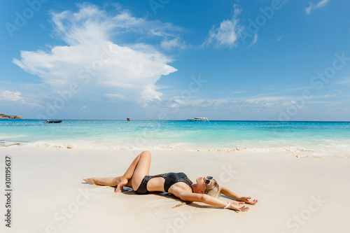 Blonde young woman in black swimsuit lying on side at beach. Beautiful tanned girl lying on sand. Attractive girl wearing glamour swimwear on tropical beach while sunbathing