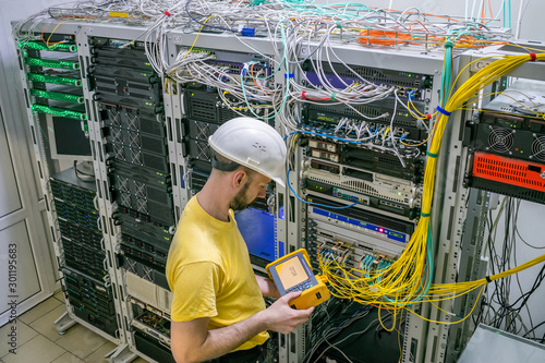 The engineer in a white helmet measures the level of the optical signal in the server room of the data center. A technician diagnoses a problem area in a telecommunication network.