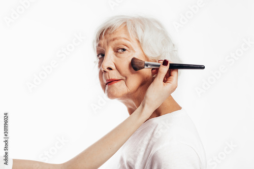 Serious old female model look at camera without smile. Woman with grey hair and red lips waiting. Young hand hold make-up brush and touch face skin. Isolated over white background.