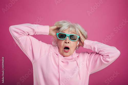 Terrified and scared old woman hold hands in hair and look straight through sunglasses. Wear pink hoody. Emotional stressed. Isolated over pink background.