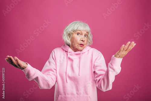 Emotional confused senior model in pink sweater stand and pose on camera. Hold hands aside of body. Wonder alone. Isolated over pink background.