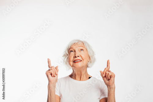 Cheerful, happy senior woman point up and look as well. Stand alone and pose on camera. Attractive female elder with grey hair. Isolated over white background.