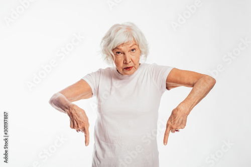 Confident old woman with wrinkles on face point down and look straight on camera. Calm peaceful elder model in white shirt stand alone. Stubborn and strong senior woman. Isolated on white background.