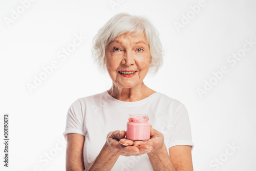 Picture of old woman with grey hair holding pink jar of face cream in hands. Look straight and smile. Anti age care and treatment at home. Isolated over white background. photo