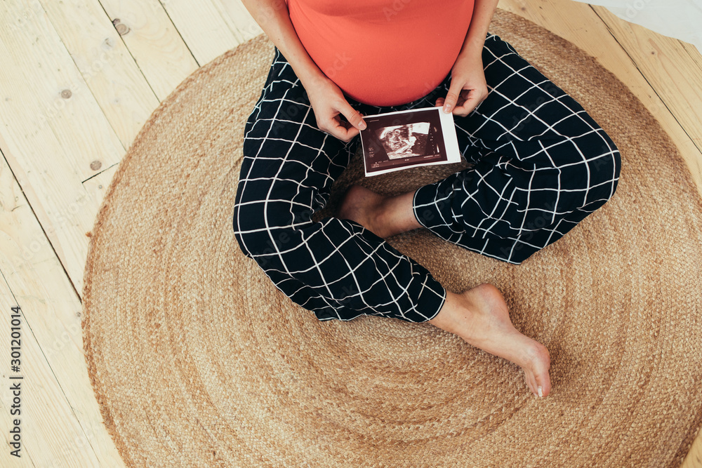 Cut view of barefeet young woman sitting on floor alone and hold ultrasound picture of her future baby. Single woman prepare to bacome mother. Maternity time soon.