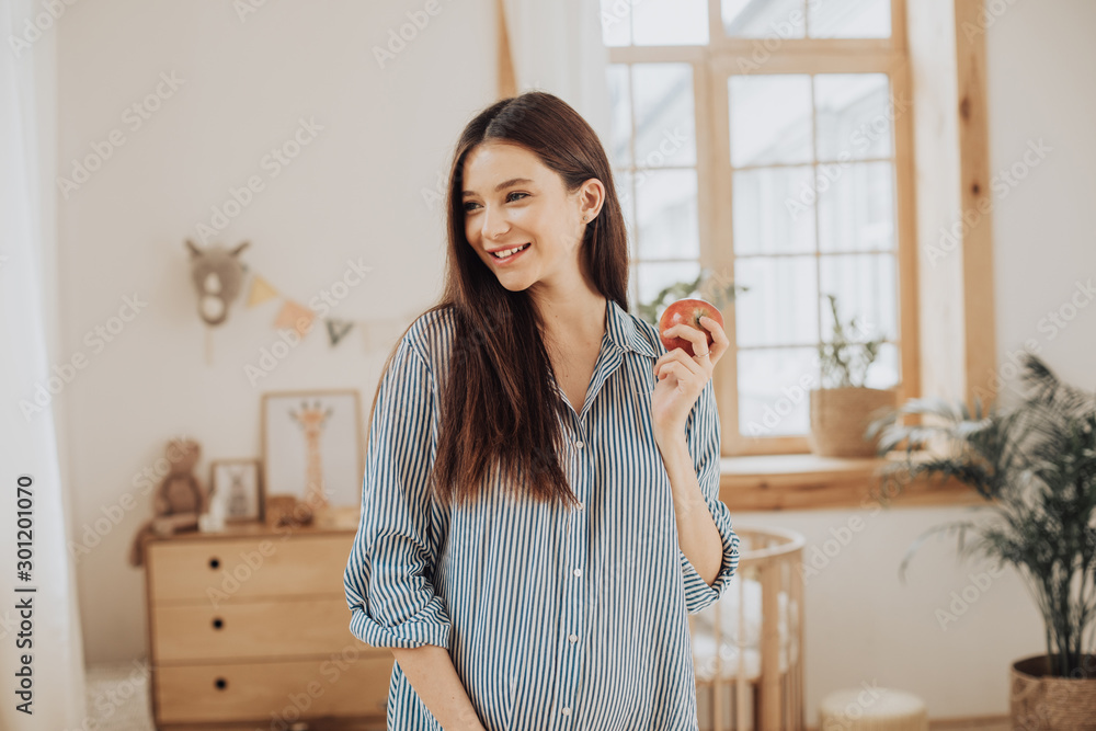 Healthy good-looking young pregnant woman stand in room alone and smile. Hold apple in hand. Wear beautiful blue dress. Waiting for child birth.