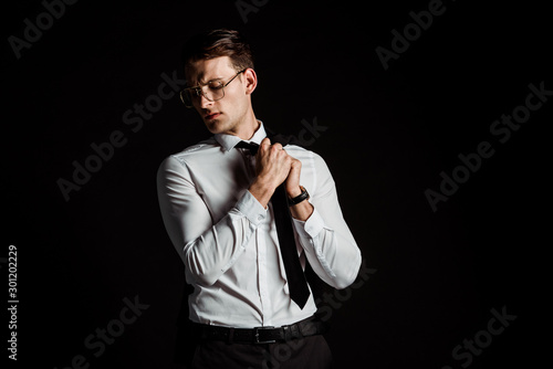 handsome businessman in glasses touching tie isolated on black