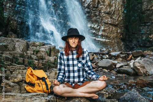 Fototapeta Naklejka Na Ścianę i Meble -  Beautiful girl with red hair in a hat and shirt meditating on rocks in a lotus position against a waterfall. Space for your text message or promotional content