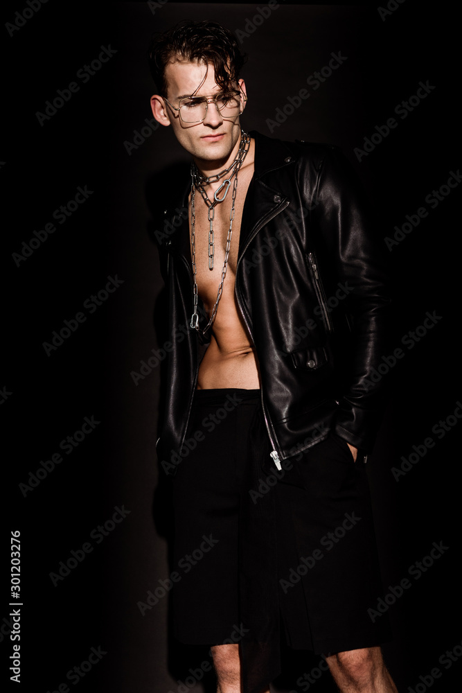 sexy man in glasses and leather jacket standing with hands in pockets on black