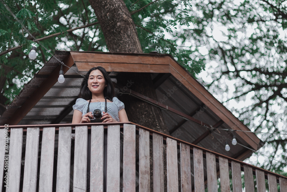 Fototapeta premium Beautiful Asian woman with film camera standing on wooden cabin which built on tree