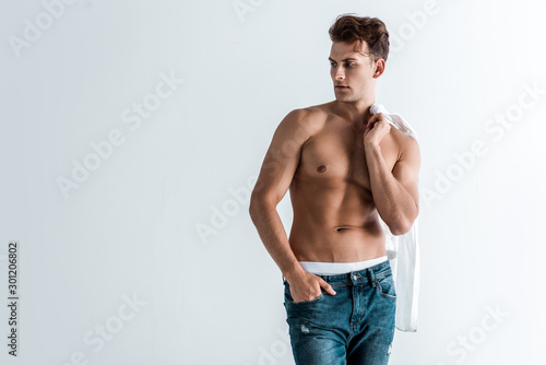 muscular young man in denim jeans standing with hand in pocket and holding shirt isolated on white