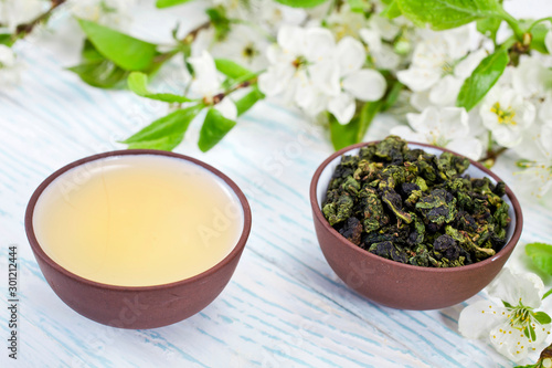 Green tea and dried leaves of green oolong tieguanyin tea in a ceramic cups with branches of blossoming apple tree on a white wooden background.