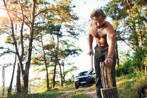 Woodsman with an axe cutting wood. Handsome shirtless man with muscular body type is in the forest at daytime