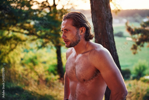 Serious and confident look. Handsome shirtless man with muscular body type is in the forest at daytime