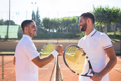 Side view of two tennis players with rackets shaking hands and smiling before tennis match